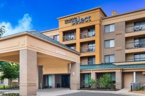 Exterior view of a Sonesta Select hotel with a covered entrance, multi-story façade featuring guest room balconies, and landscaped grounds under a blue sky.